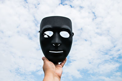 Cropped hand of person holding black mask against cloudy sky