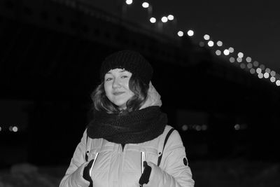 Portrait of happy young woman in warm clothing standing against illuminated bridge