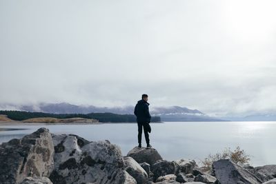 Rear view of man standing on rock by lake against sky