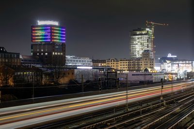 Light trails on road by buildings against sky at night