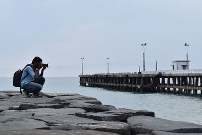 Full length side view of man crouching while photographing on groyne at beach