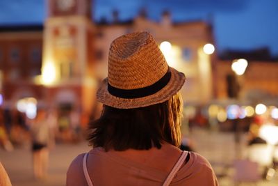 Rear view of woman in hat standing against illuminated city