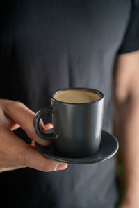 A man's hand holds a cup of hot espresso coffee against the background of a black t-shirt.