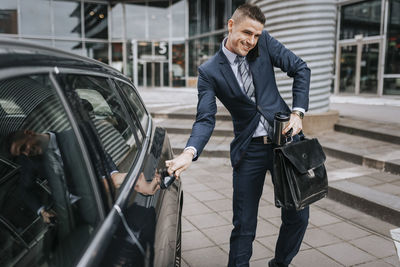 Businessman talking on smart phone while opening car door near office building