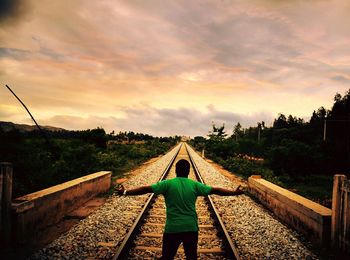Rear view of man standing with arms outstretched on railroad tracks during sunset