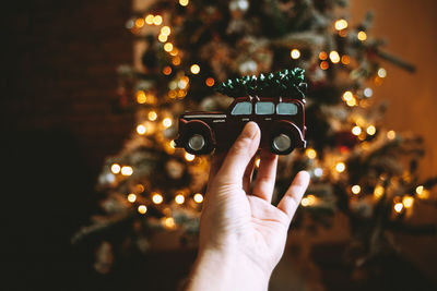 Cropped hand of woman holding toy car against illuminated christmas tree