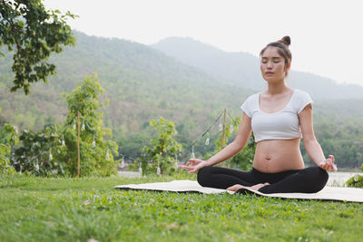 Full length of pregnant young woman meditating while sitting on mat over grass
