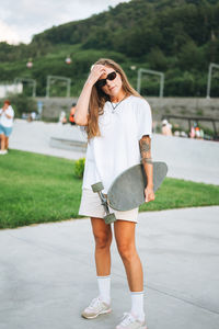 Slim young woman with long blonde hair in light sports clothes with longboard in outdoor skatepark 