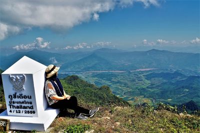 Side view of woman sitting by sign on cliff against mountains