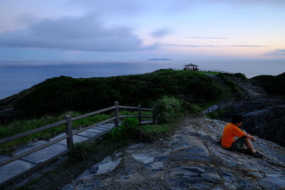 Side view of man sitting on mountain by sea against sky during sunset