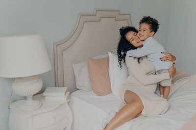 Cheerful mother embracing son sitting on bed at home