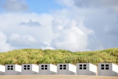 Beach huts by grassy hill against cloudy sky