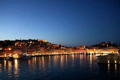 Illuminated buildings by river against blue sky