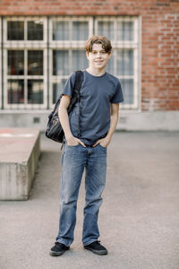 Portrait of male teenage student standing with hands in pockets in high school campus