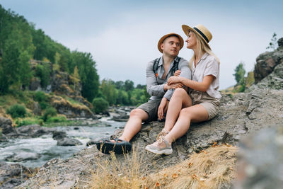 Full length of couple sitting on rock by river in forest