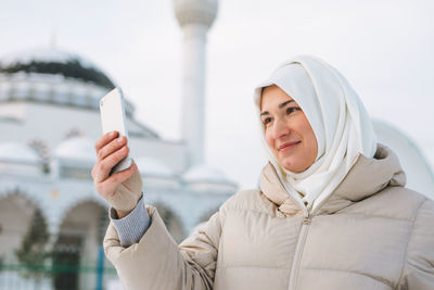 Beautiful smiling young muslim woman in headscarf in clothing takes selfie against mosque in winter 