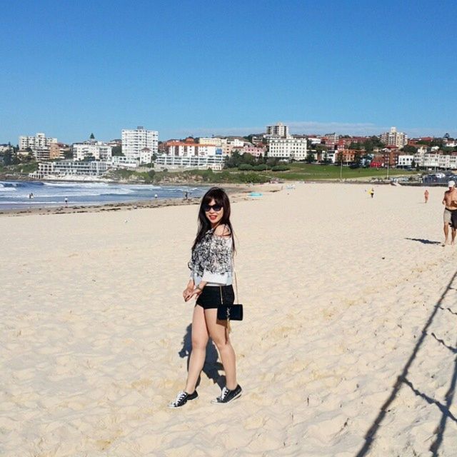 beach, building exterior, clear sky, architecture, lifestyles, water, full length, built structure, leisure activity, casual clothing, young adult, sand, person, blue, shore, sea, city, sunlight