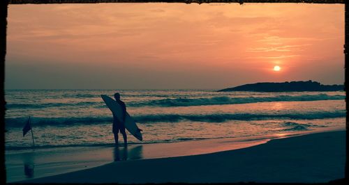 Silhouette man holding surfboard and standing on shore against sky during sunset