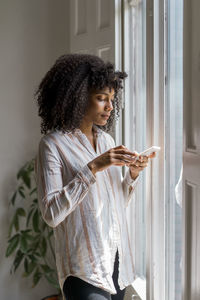 Beautiful female entrepreneur using smart phone while standing near window in office