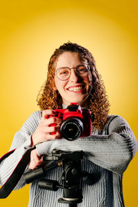 Portrait of young woman with camera against yellow background