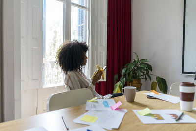 Young afro female entrepreneur using smart phone while having banana during coffee break at office