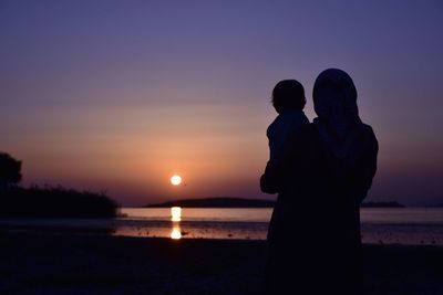 Silhouette mother with child at beach against sky during sunset