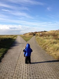 Rear view of boy waring winter coat standing on footpath against sky