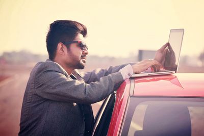 Side view of young man using laptop on car against sky