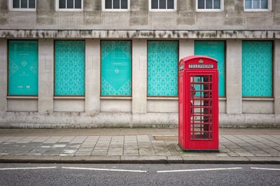 Red telephone booth on sidewalk by building in city