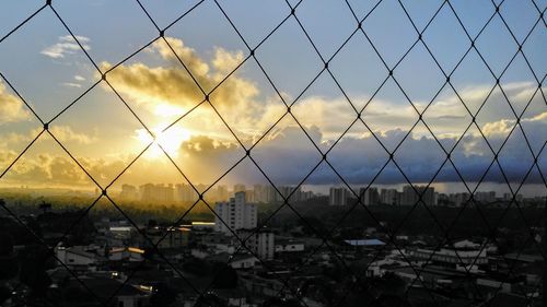 Silhouette buildings seen through chainlink fence against sky during sunset