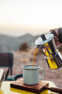 Crop anonymous hiker pouring hot brewed coffee from moka pot into tin mug for breakfast in nature during trekking trip