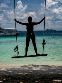 Rear view of young woman standing on wooden swing by the beach