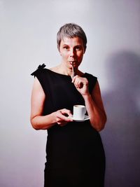 Mature woman holding coffee cup while standing against wall