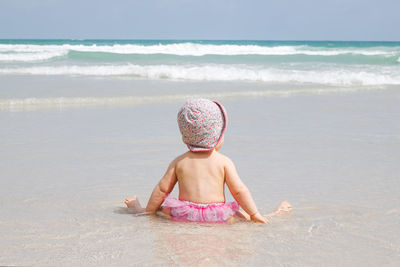 Little baby girl is sitting at sandy beach and looking at the blue sea with waves.