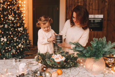 Happy mom and daughter in sweaters prepare a festive cake for christmas holiday in kitchen at home