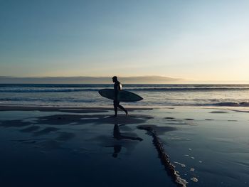 Full length of surfer walking at beach against clear sky during sunset