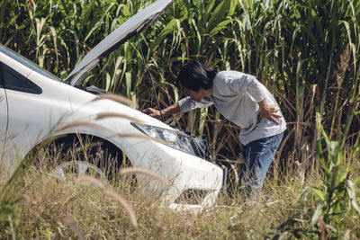 Side view of man standing by damaged car against plants