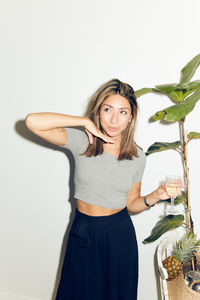 Smiling young woman holding drink while standing by plant against white wall at home