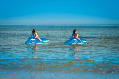 Siblings on inflatable rafts in sea against blue sky