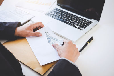 Midsection of businessman with paper currency in envelope by laptop on desk