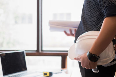 Midsection of engineer with hardhat standing by laptop on desk at office