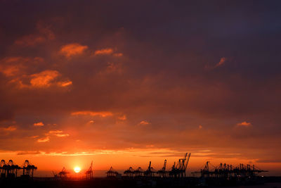 Silhouette cranes at harbor against sky during sunset