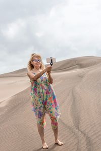 Vertical photo of a woman with sunglasses using a portable tripod to take a selfie in a dune