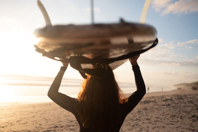 Rear view of mature woman carrying surfboard on head at beach