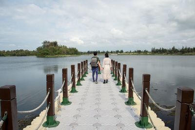 Rear view of couple holding hands while walking on pier over lake against sky
