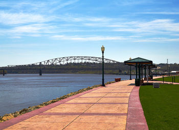 Walkway by river against sky on sunny day