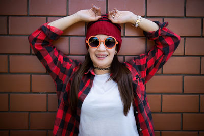 Portrait of smiling woman in sunglasses and knit hat standing against brick wall