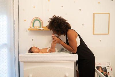 Side view of tender african american mother caressing adorable toddler lying in diaper on changing table at home