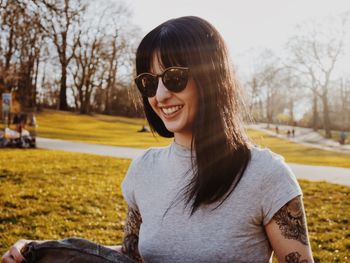 Smiling young woman with tattoos wearing sunglasses at park on sunny day