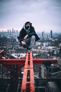 Young man looking away while sitting on metal railing against sky